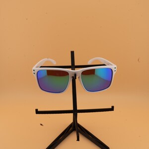 May include: White-framed sunglasses with reflective blue and green lenses. The sunglasses are displayed on a black stand against a light orange background. The sunglasses have a square shape.