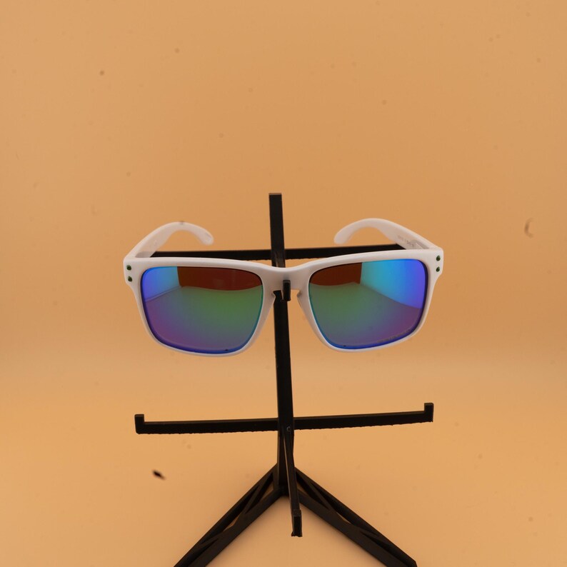 May include: White-framed sunglasses with reflective blue and green lenses. The sunglasses are displayed on a black stand against a light orange background. The sunglasses have a square shape.