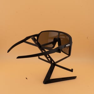 May include: Black sunglasses with brown lenses are displayed on a black stand. The sunglasses have a sleek, modern design with a prominent frame and curved arms. The lenses are tinted brown, offering a contrast to the black frame.