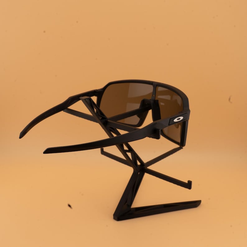 May include: Black sunglasses with brown lenses are displayed on a black stand. The sunglasses have a sleek, modern design with a prominent frame and curved arms. The lenses are tinted brown, offering a contrast to the black frame.