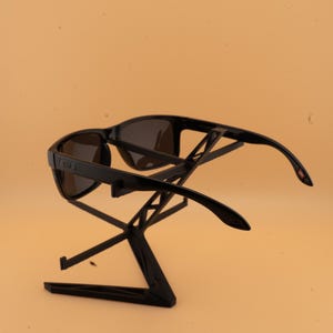 May include: Black sunglasses with dark lenses are displayed on a black stand. The sunglasses have a rectangular frame and curved arms. The stand is a geometric design, providing a modern display for the eyewear.
