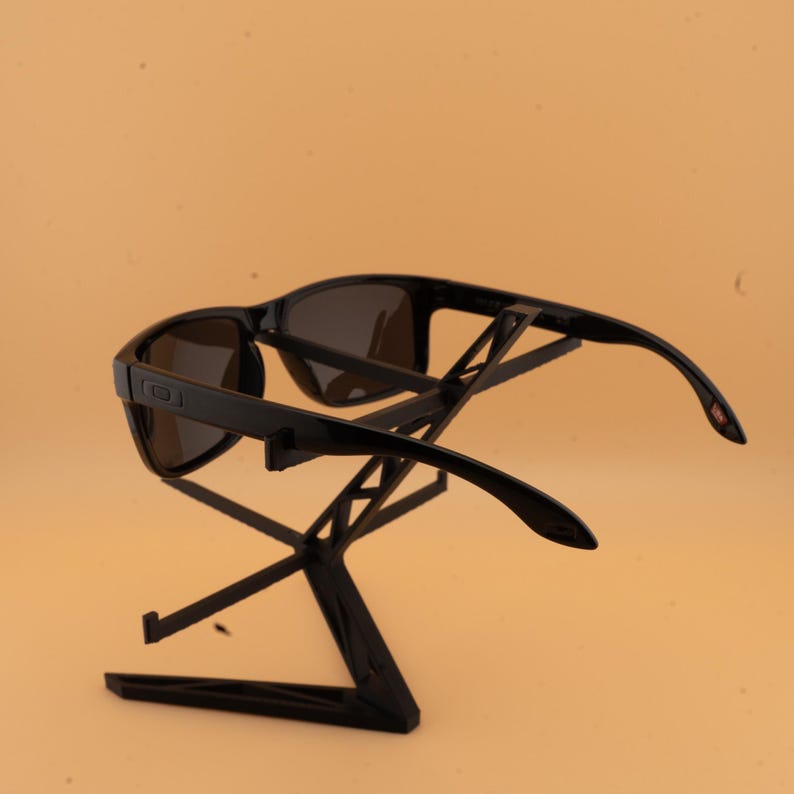 May include: Black sunglasses with dark lenses are displayed on a black stand. The sunglasses have a rectangular frame and curved arms. The stand is a geometric design, providing a modern display for the eyewear.
