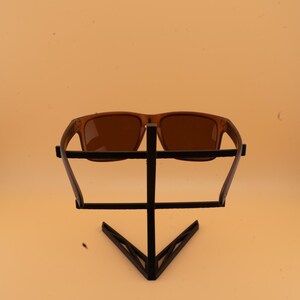 May include: Brown sunglasses with a rectangular frame are displayed on a black metal stand. The sunglasses have a classic design with a solid brown tint. The stand has a geometric design.