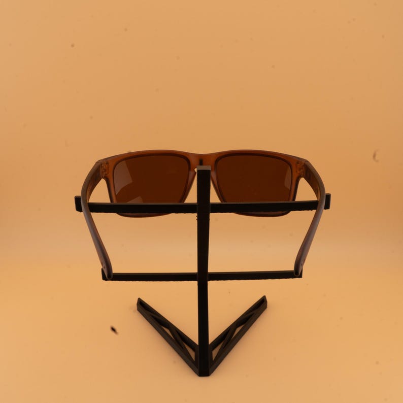 May include: Brown sunglasses with a rectangular frame are displayed on a black metal stand. The sunglasses have a classic design with a solid brown tint. The stand has a geometric design.