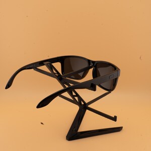 May include: Black sunglasses with dark lenses are displayed on a black stand against a light orange background. The sunglasses have a rectangular frame and curved arms. The stand is a geometric design.