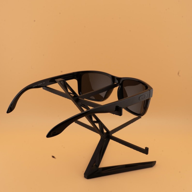 May include: Black sunglasses with dark lenses are displayed on a black stand against a light orange background. The sunglasses have a rectangular frame and curved arms. The stand is a geometric design.