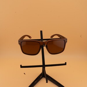 May include: Brown sunglasses with a square frame and tinted lenses are displayed on a black metal stand. The sunglasses have a classic design, suitable for various styles. The background is a solid, light orange color.