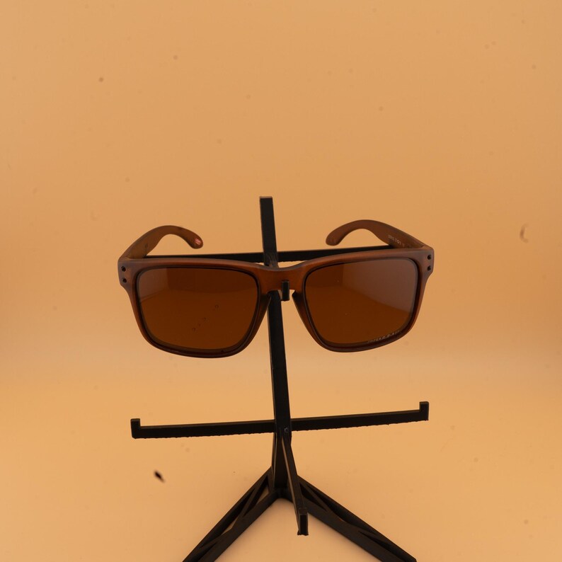 May include: Brown sunglasses with a square frame and tinted lenses are displayed on a black metal stand. The sunglasses have a classic design, suitable for various styles. The background is a solid, light orange color.