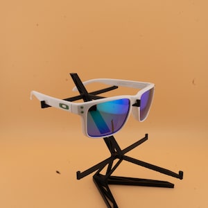 May include: White-framed sunglasses with blue-green reflective lenses. The sunglasses are displayed on a black stand against a light orange background. The frames have a rectangular shape and a small green logo on the side.