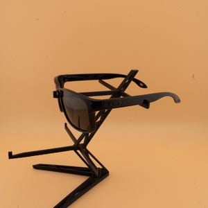 May include: Black sunglasses with dark lenses are displayed on a black geometric stand. The sunglasses have a rectangular frame and curved temples. The stand has a modern, angular design, providing a stylish presentation for the eyewear.