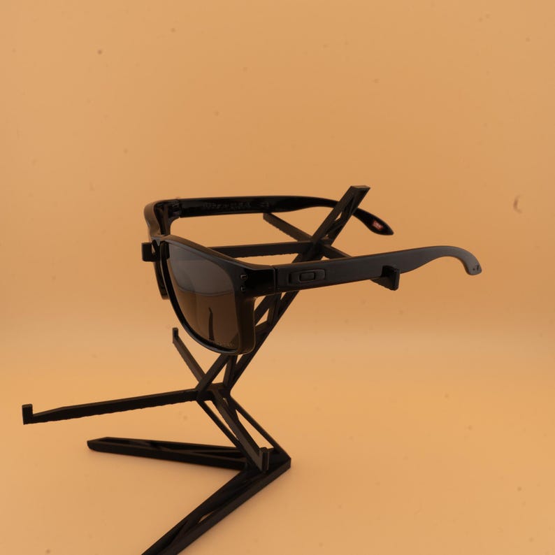 May include: Black sunglasses with dark lenses are displayed on a black geometric stand. The sunglasses have a rectangular frame and curved temples. The stand has a modern, angular design, providing a stylish presentation for the eyewear.