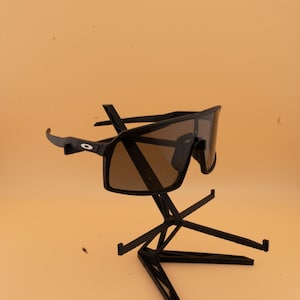 May include: Black sunglasses with brown lenses are displayed on a black stand. The sunglasses have a sporty, wraparound design. The stand has a geometric, multi-level design, and the background is a solid, light orange color.