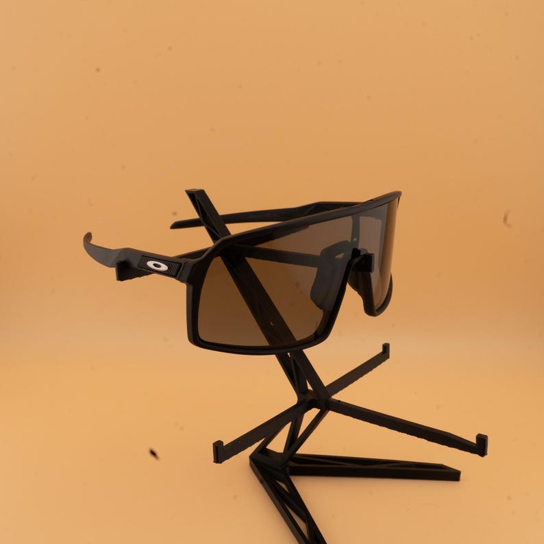 May include: Black sunglasses with brown lenses are displayed on a black stand. The sunglasses have a sporty, wraparound design. The stand has a geometric, multi-level design, and the background is a solid, light orange color.