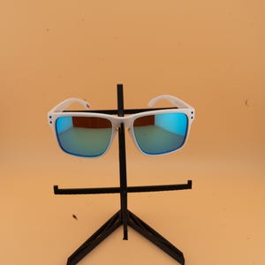 Puede incluir: Gafas de sol con montura blanca y lentes espejadas azules expuestas en un soporte negro. Las gafas de sol tienen forma cuadrada y un dise&ntilde;o moderno. Las lentes reflejan un tono azul, y las monturas son de color blanco.