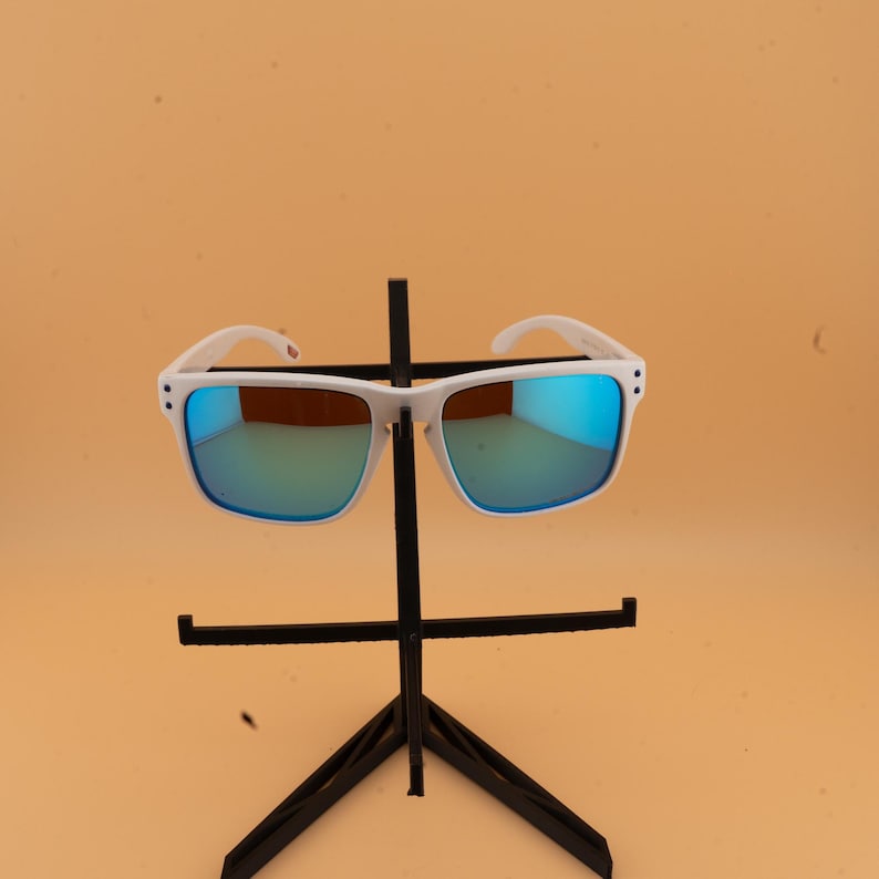 Puede incluir: Gafas de sol con montura blanca y lentes espejadas azules expuestas en un soporte negro. Las gafas de sol tienen forma cuadrada y un dise&ntilde;o moderno. Las lentes reflejan un tono azul, y las monturas son de color blanco.
