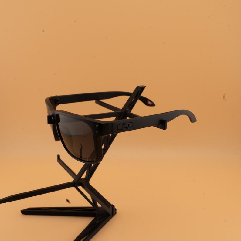 May include: Black sunglasses with tinted lenses are presented on a black stand against a light orange backdrop. The sunglasses feature a rectangular frame and curved arms. The stand has a geometric design.
