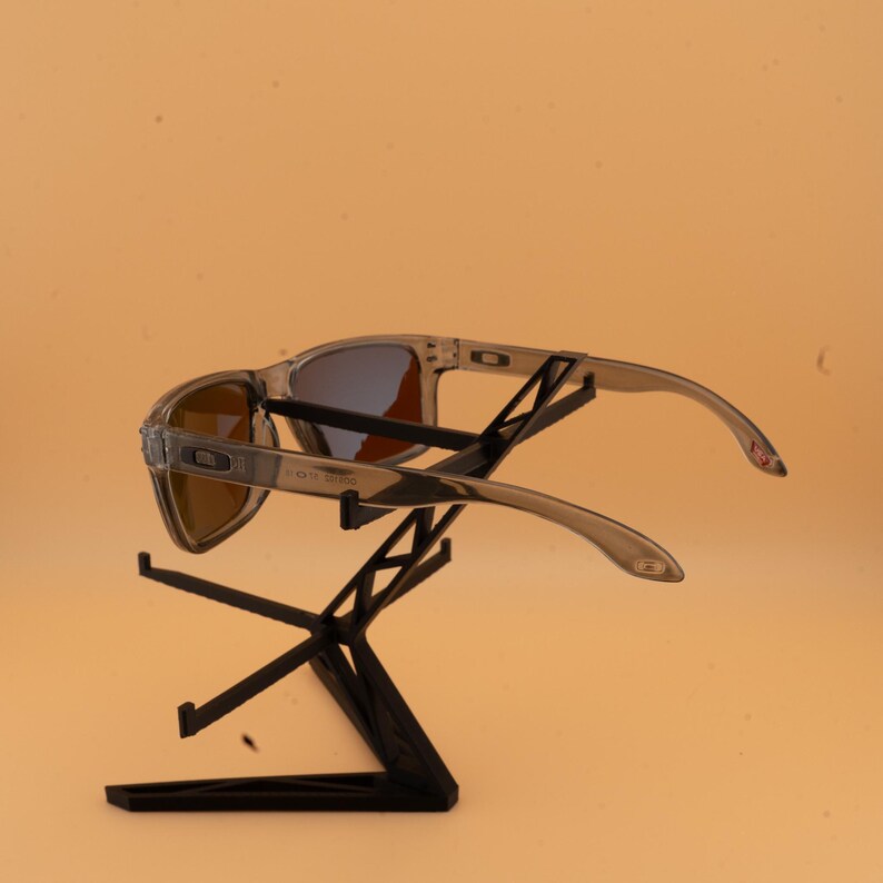 Puede incluir: Gafas de sol con montura transparente y lentes marrones se exhiben en un soporte negro. Las gafas de sol tienen un dise&ntilde;o deportivo con una montura y patillas curvas. Las lentes tienen un tinte degradado y las patillas tienen un acento rojo.