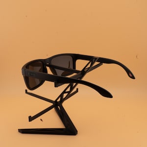 May include: Black sunglasses with dark lenses are presented on a black stand. The sunglasses feature a rectangular frame and curved arms. The stand has a geometric design, offering a modern look. The background is a solid, light orange colour.