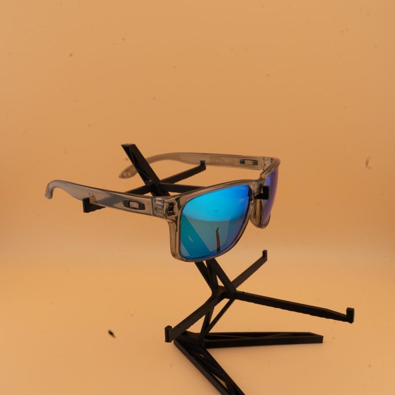 Puede incluir: Gafas de sol grises con lentes azules reflectantes se exhiben en un soporte negro. Las gafas de sol tienen una montura rectangular y un aspecto claro y transl&uacute;cido. Las lentes reflejan un color azul vibrante.