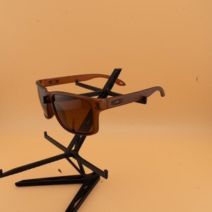 May include: Brown sunglasses with a square frame and dark lenses. The sunglasses have a light brown frame and are displayed on a black metal stand against a light orange background. The sunglasses are a stylish accessory.