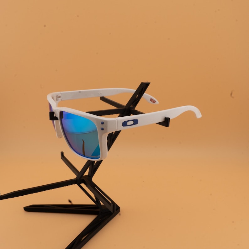 Puede incluir: Gafas de sol blancas con lentes de espejo azules. Las gafas tienen una montura blanca con un logotipo azul en las patillas. Las lentes reflejan un tono azul. Las gafas se exhiben en un soporte negro.