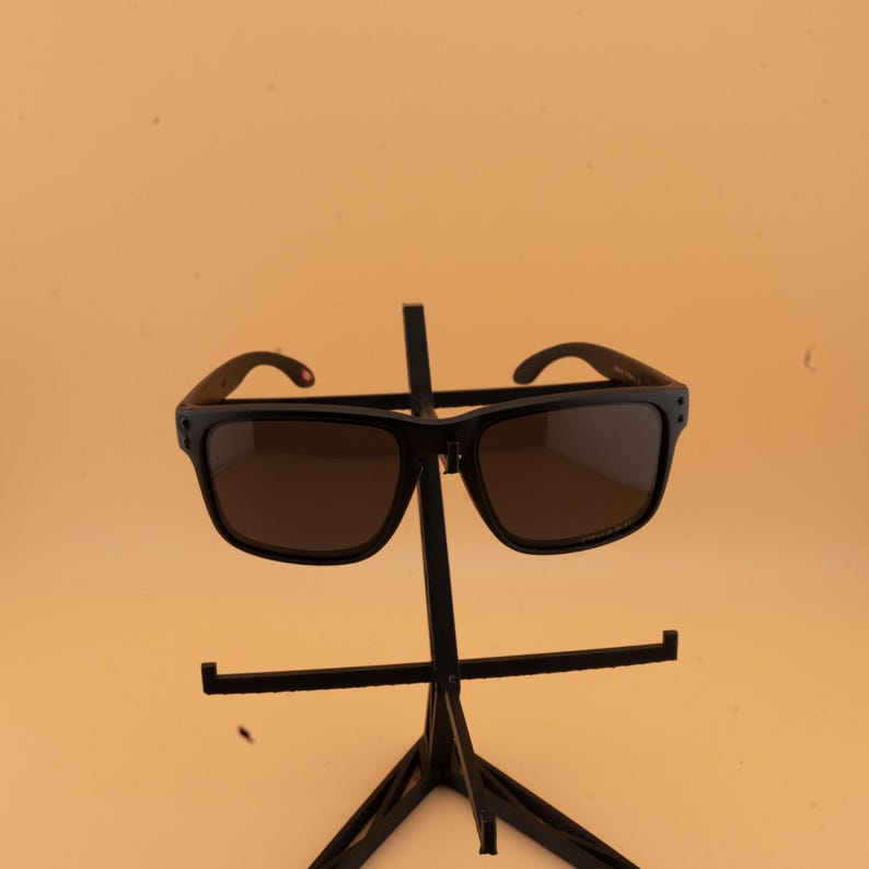 May include: Black sunglasses with dark lenses. The sunglasses have a square frame and are displayed on a black stand. The arms of the sunglasses have a small red accent. The background is a solid, light orange colour.