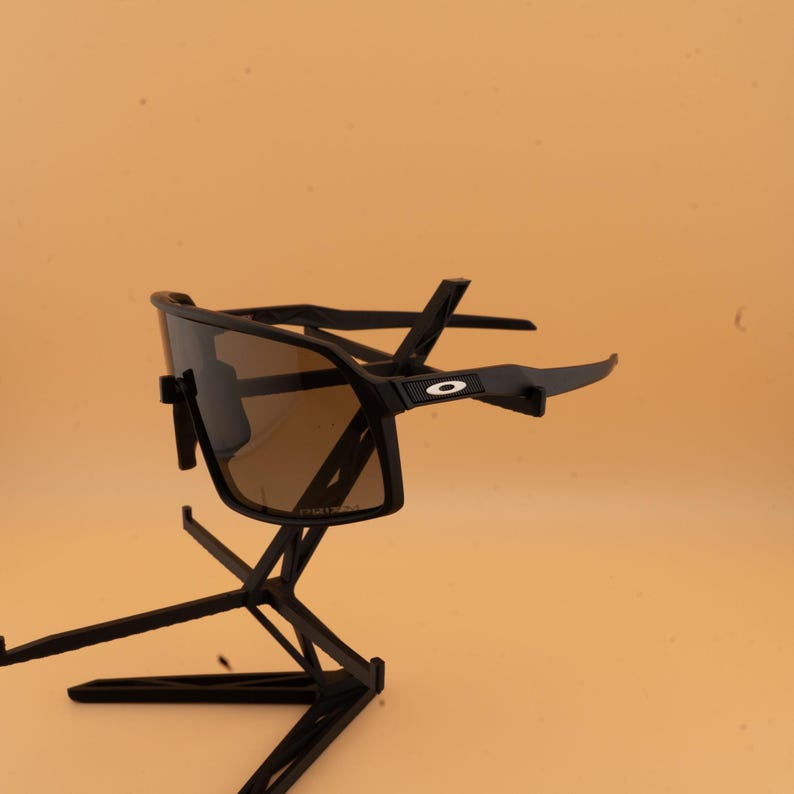 May include: Black sunglasses with brown lenses are displayed on a black stand. The sunglasses have a sporty design with a thick frame and a small white circle detail on the arm. The stand has a geometric design.