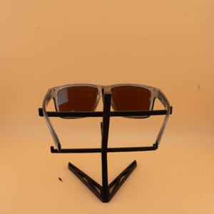 Puede incluir: Gafas de sol con montura transparente y lentes marrones expuestas en un soporte de metal negro. Las gafas de sol tienen forma rectangular y est&aacute;n sobre un fondo naranja claro.