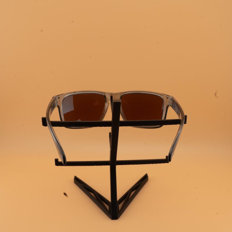 Puede incluir: Gafas de sol con montura transparente y lentes marrones expuestas en un soporte de metal negro. Las gafas de sol tienen forma rectangular y est&aacute;n sobre un fondo naranja claro.