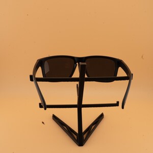 May include: Black sunglasses with dark lenses are displayed on a black metal stand. The sunglasses have a rectangular frame. The stand has a geometric design with a triangular base and horizontal bars.