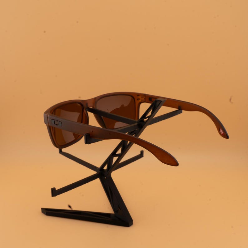 May include: Brown sunglasses with a rectangular frame and tinted lenses are displayed on a black metal stand. The sunglasses have a sleek, modern design, and the stand provides an elevated presentation against a light orange background.