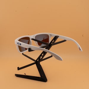 Puede incluir: Gafas de sol blancas con lentes marrones expuestas en un soporte negro. Las gafas tienen una montura rectangular y un peque&ntilde;o logotipo azul y blanco en el lateral. Las patillas se curvan ligeramente.