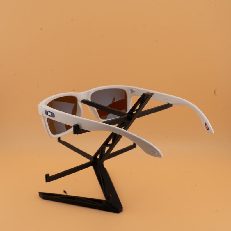Puede incluir: Gafas de sol blancas con lentes marrones expuestas en un soporte negro. Las gafas tienen una montura rectangular y un peque&ntilde;o logotipo azul y blanco en el lateral. Las patillas se curvan ligeramente.