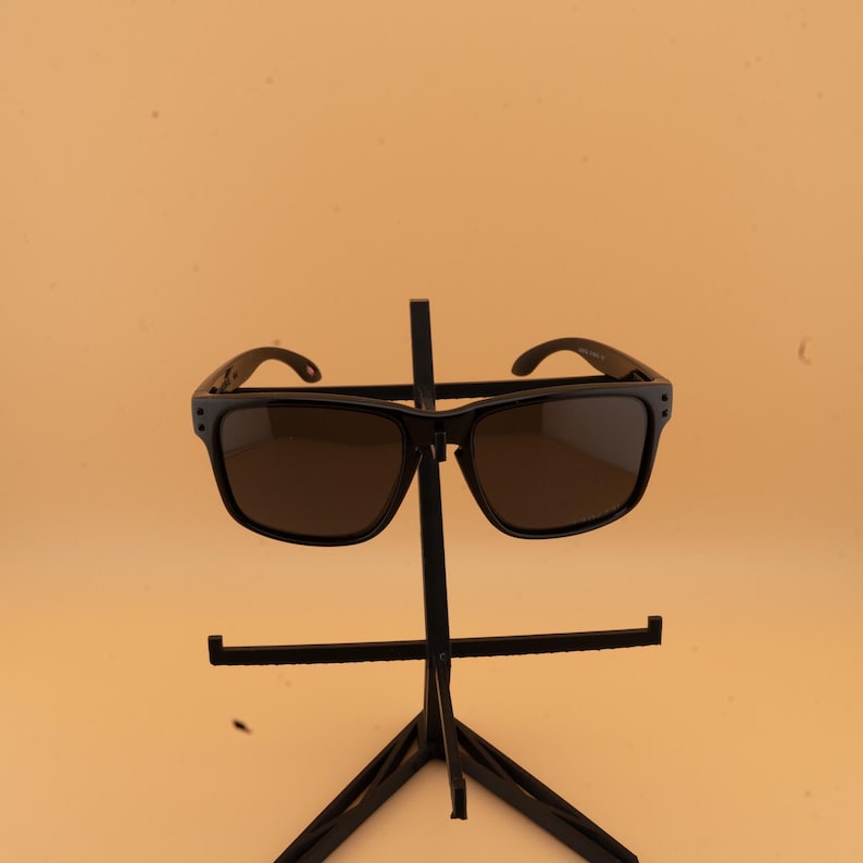 May include: Black sunglasses with dark lenses are displayed on a black stand. The sunglasses have a square frame and curved arms. The stand has a three-legged base and a vertical support with horizontal bars to hold the glasses.