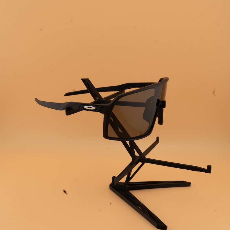 May include: Black sunglasses with a dark lens are displayed on a black stand. The sunglasses have a sporty design with a rectangular frame and curved arms. The stand has a geometric design.