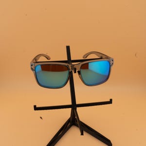 Puede incluir: Gafas de sol de montura cuadrada con montura gris transl&uacute;cida y lentes espejadas azules. Las gafas de sol se exhiben en un soporte negro sobre un fondo naranja claro. Las lentes reflejan azul y verde.