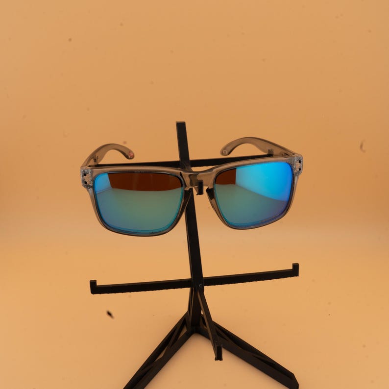 Puede incluir: Gafas de sol de montura cuadrada con montura gris transl&uacute;cida y lentes espejadas azules. Las gafas de sol se exhiben en un soporte negro sobre un fondo naranja claro. Las lentes reflejan azul y verde.