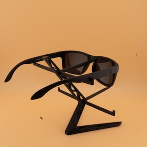 May include: Black sunglasses with dark lenses are displayed on a black stand. The sunglasses have a rectangular frame and curved arms. The stand is a geometric design with multiple intersecting lines.