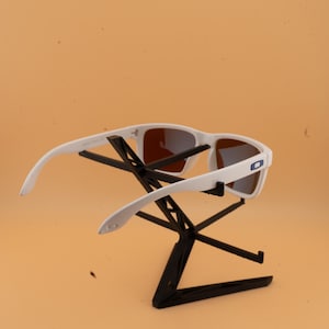 Puede incluir: Gafas de sol blancas con lentes marrones se exhiben en un soporte negro. Las gafas de sol tienen un dise&ntilde;o moderno con montura blanca y un logotipo azul en el lateral. El soporte tiene forma geom&eacute;trica.