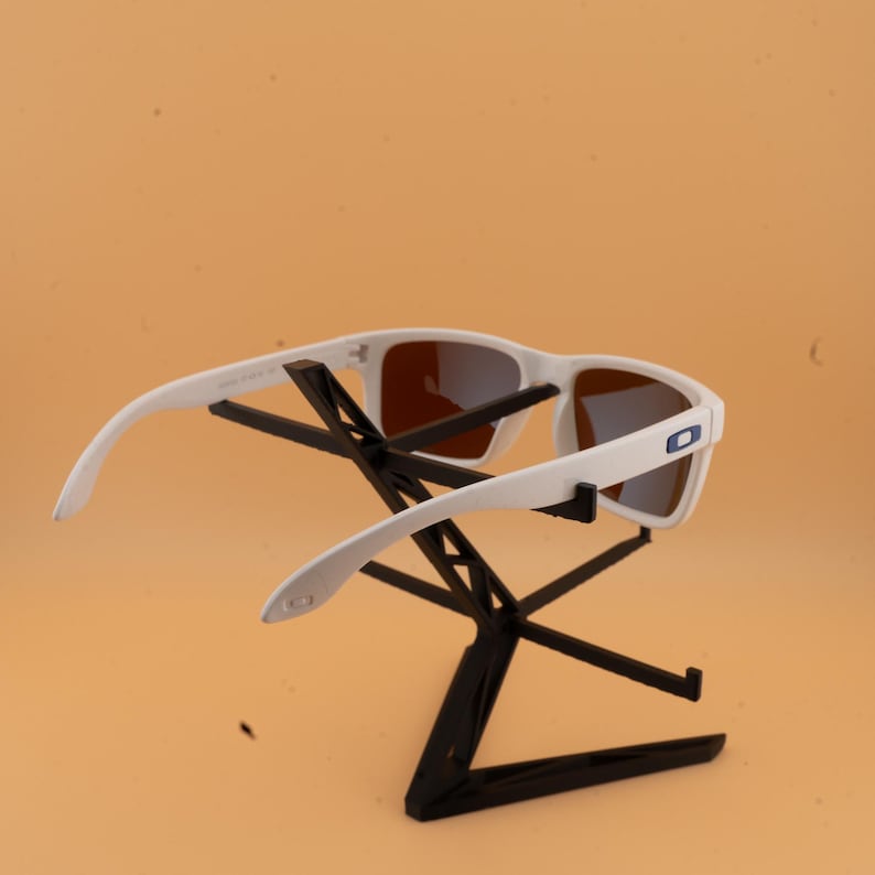 Puede incluir: Gafas de sol blancas con lentes marrones se exhiben en un soporte negro. Las gafas de sol tienen un dise&ntilde;o moderno con montura blanca y un logotipo azul en el lateral. El soporte tiene forma geom&eacute;trica.