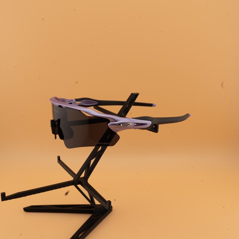 以下が含まれることがあります： 紫色のフレームと濃い色のレンズを備えたスポーツサングラス。黒いスタンドに置かれ、明るいオレンジ色の背景に配置されています。 スタイリッシュでモダンなデザインです。