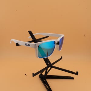 Puede incluir: Gafas de sol blancas con lentes azules espejadas. Las gafas se exhiben en un soporte negro sobre un fondo naranja claro. Las monturas tienen forma rectangular y un peque&ntilde;o logotipo en el lateral.