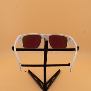 May include: White-framed sunglasses with red-tinted lenses. The sunglasses are displayed on a black metal stand against a light orange background. The arms of the sunglasses are extended.