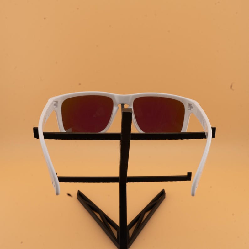 May include: White-framed sunglasses with red-tinted lenses. The sunglasses are displayed on a black metal stand against a light orange background. The arms of the sunglasses are extended.