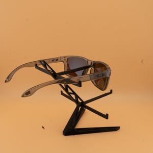 Puede incluir: Un par de gafas de sol con montura transparente y lentes azules y marrones descansa sobre un expositor negro. Las gafas de sol tienen un dise&ntilde;o moderno y el expositor est&aacute; hecho de finas l&iacute;neas que se cruzan.