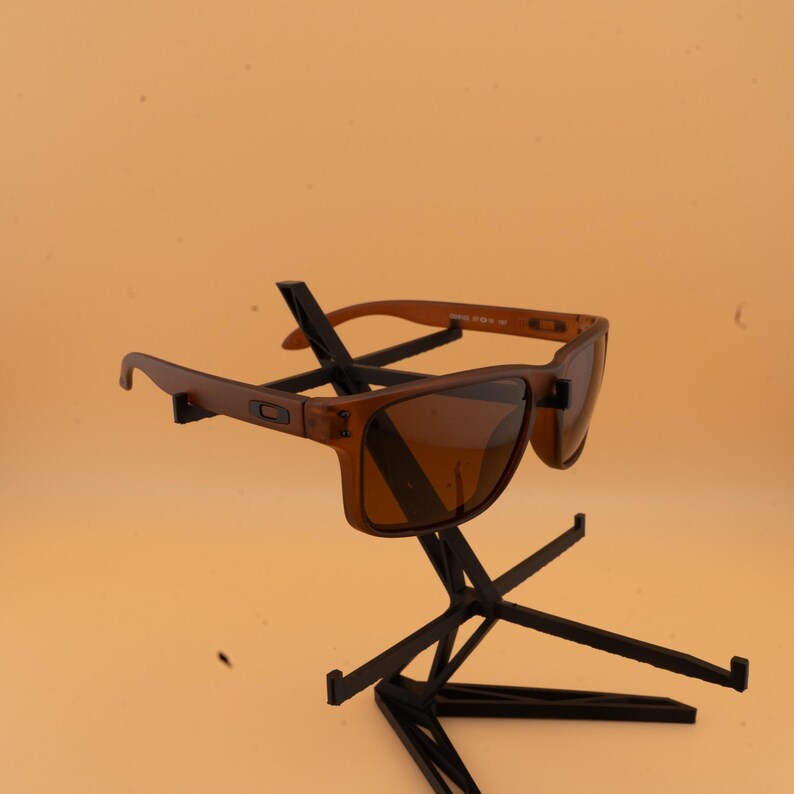 May include: Brown sunglasses with a rectangular frame and tinted lenses. The sunglasses are displayed on a black stand against a light orange background. The arms of the sunglasses are a lighter shade of brown.