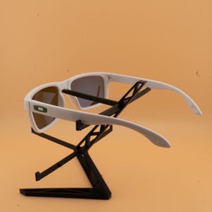 May include: White sunglasses with gray lenses are displayed on a black stand. The sunglasses have a modern design with a white frame and a green logo on the side. The stand is a geometric shape.