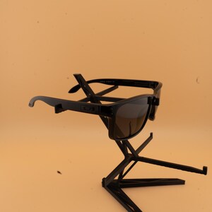 May include: Black sunglasses with dark lenses are displayed on a black stand. The sunglasses have a rectangular shape and a thick frame. The stand is a geometric design, providing a modern display for the eyewear.
