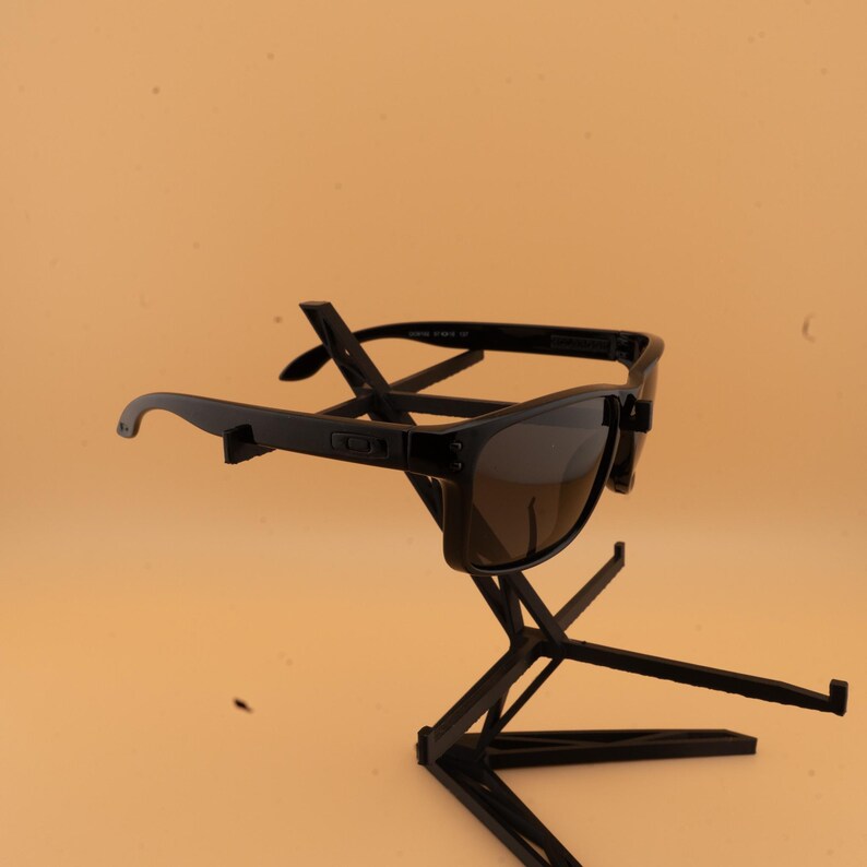 May include: Black sunglasses with dark lenses are displayed on a black stand. The sunglasses have a rectangular shape and a thick frame. The stand is a geometric design, providing a modern display for the eyewear.