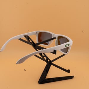 May include: White sunglasses with a rectangular frame and mirrored lenses. The sunglasses are displayed on a black stand against a light orange background. The sunglasses have a green logo on the side.